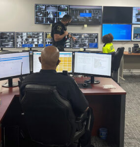 The image shows a security control room with several large monitors displaying various camera feeds
